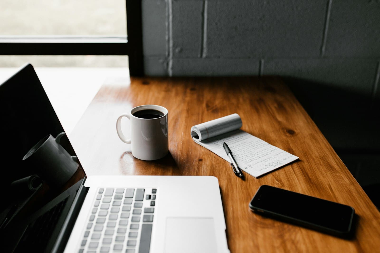 A laptop, coffee mug, notepad and phone arranged on a wooden desk.