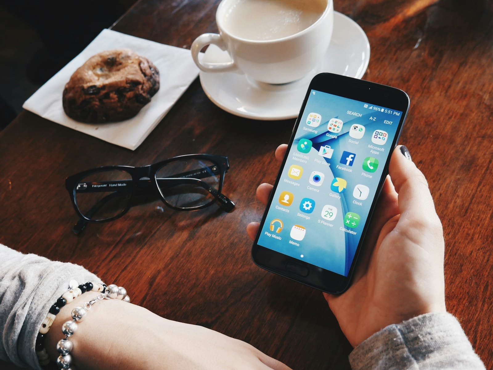 A person holding a smartphone showing a home screen of apps, at a cafe table with coffee and a biscuit.
