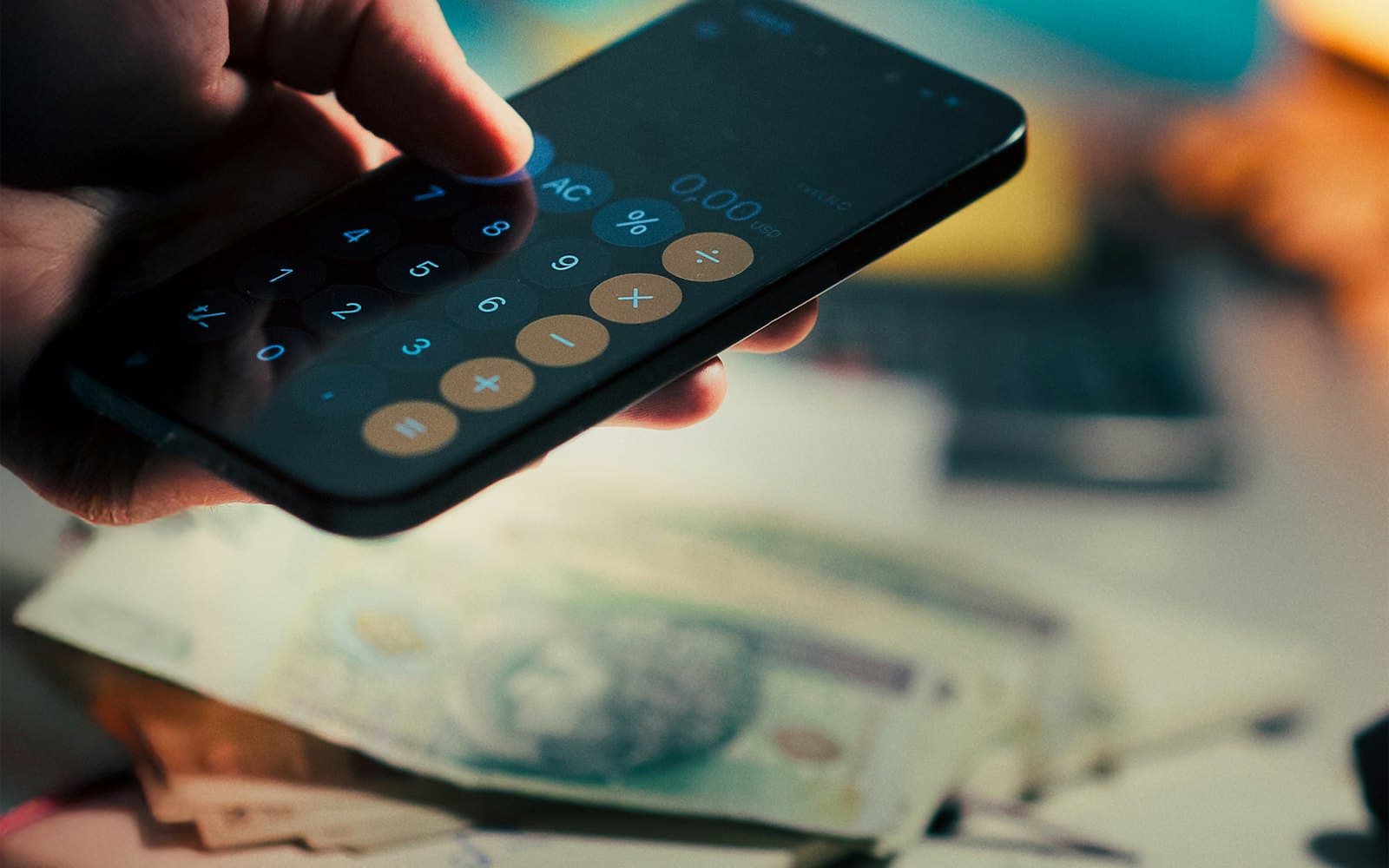 A hand using a calculator app on a smartphone above a desk with banknotes and notebooks.