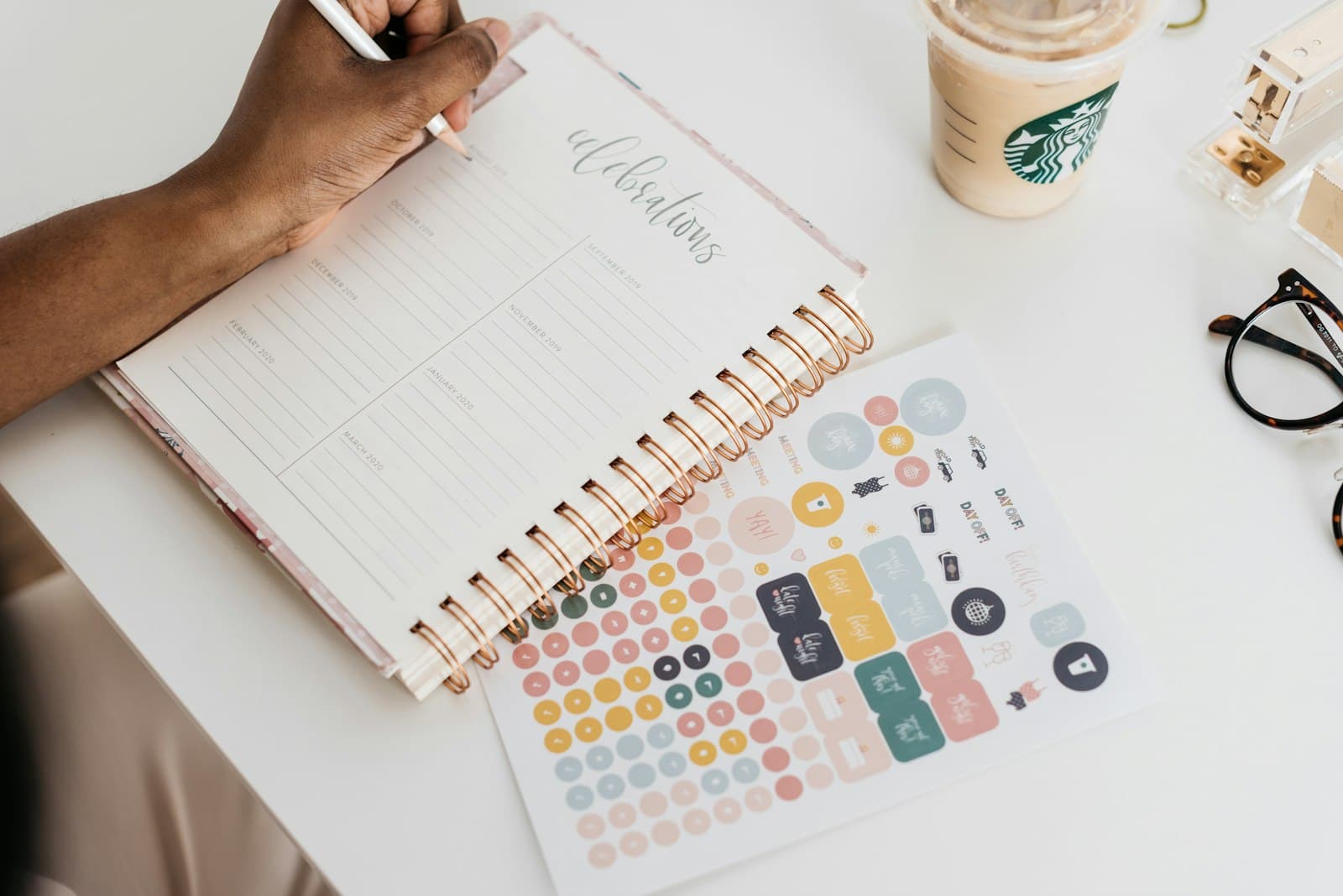 A person writing in a spiral planner at a white desk.