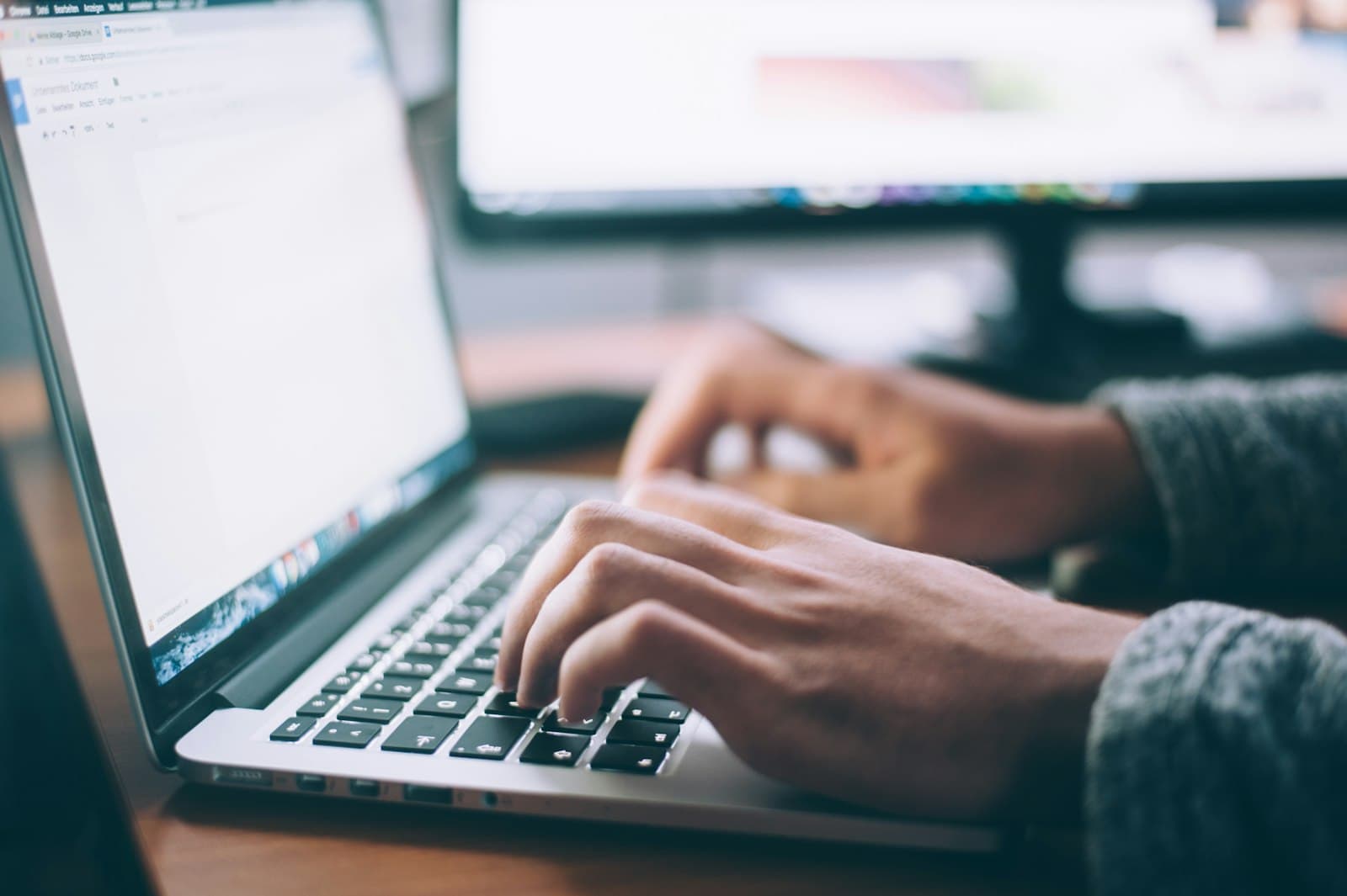 Hands typing on a laptop keyboard with a second monitor blurred in the background.
