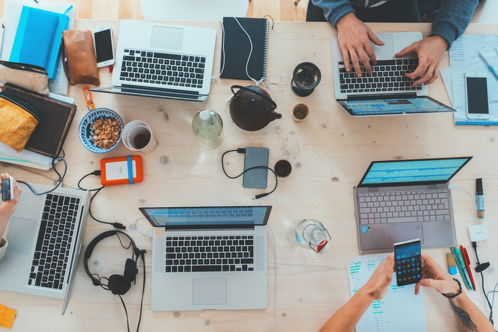 A shared table seen from above, covered with laptops, phones, headphones and notebooks.