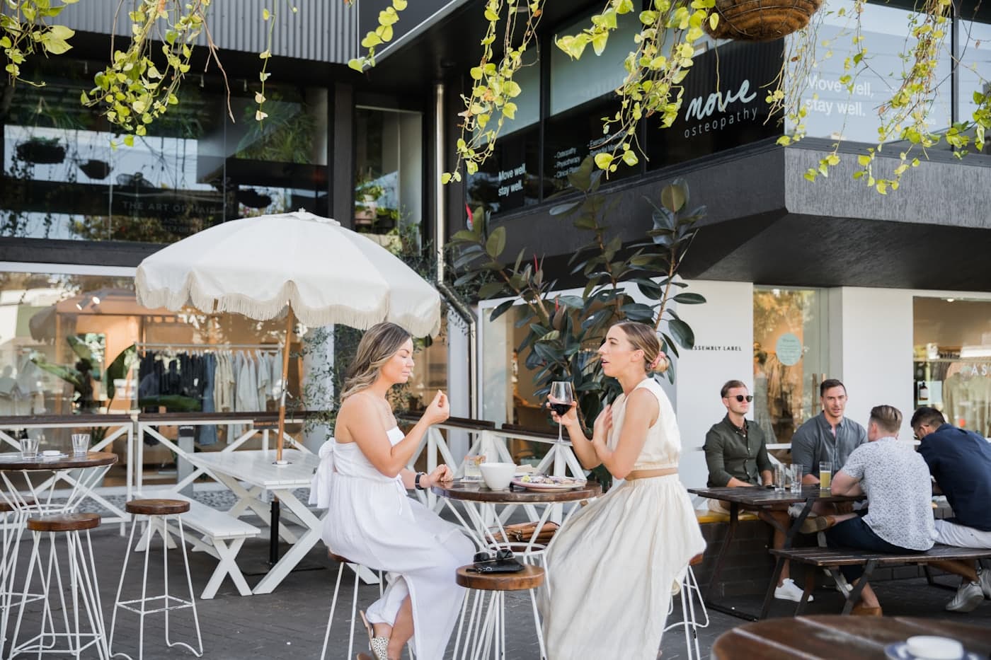 People eating and talking at outdoor cafe tables under a white umbrella on an Australian street.