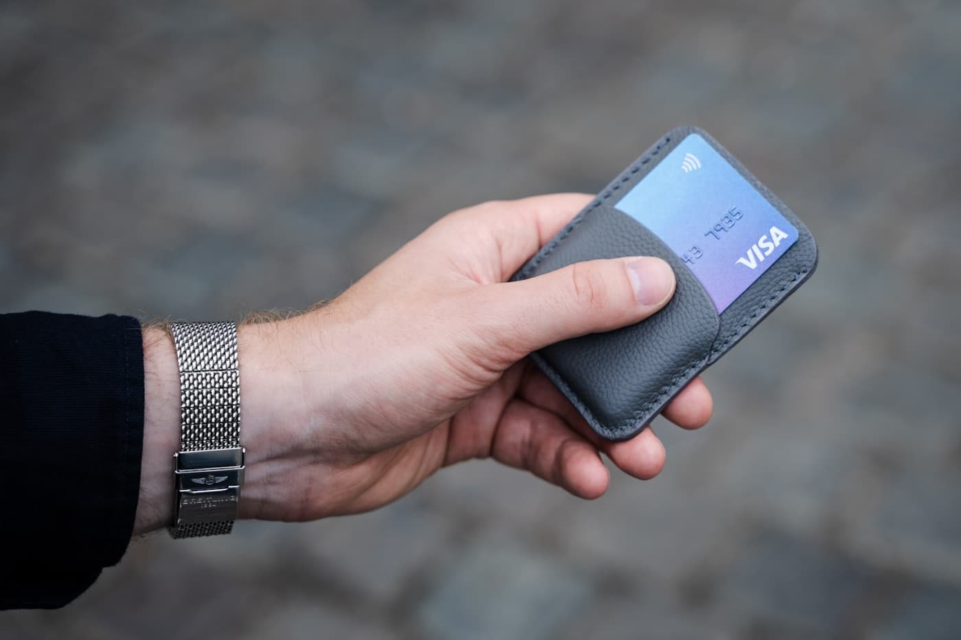 A hand holding a small grey card wallet with a contactless Visa card tucked inside.