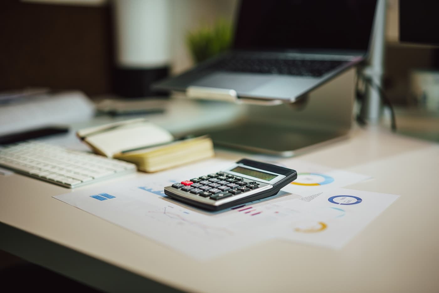 A desk with a calculator resting on printed spending charts beside a laptop.