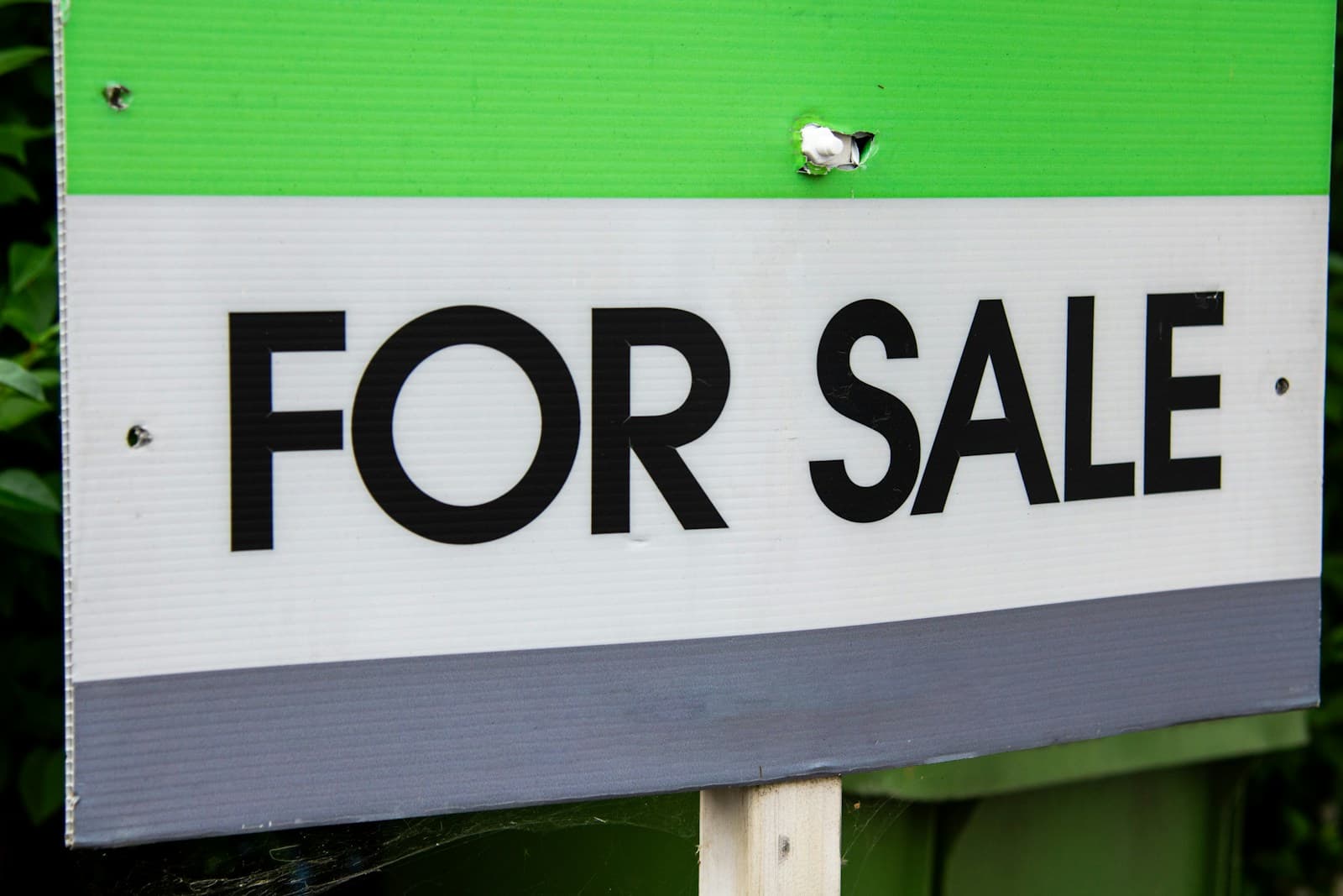 A plain green and white for sale sign in front of a hedge outside a house.