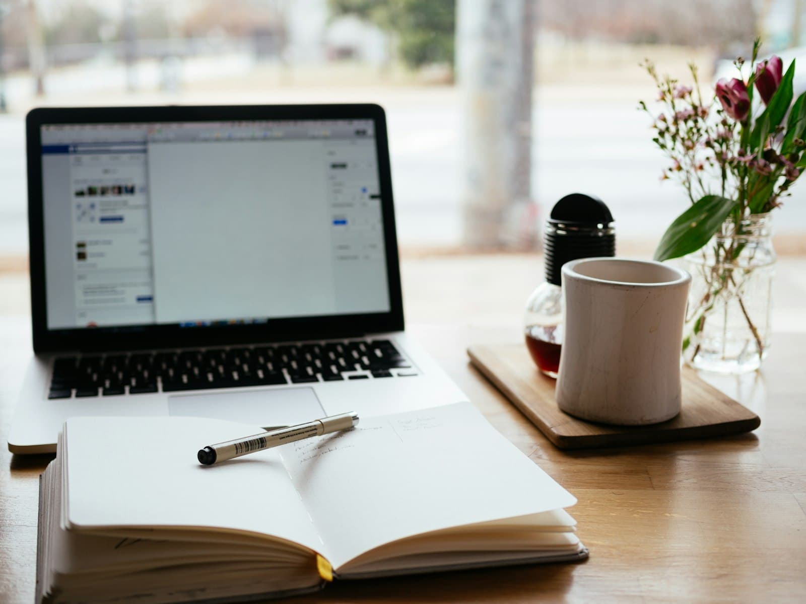 A laptop and an open notebook with a pen on a desk by a window.