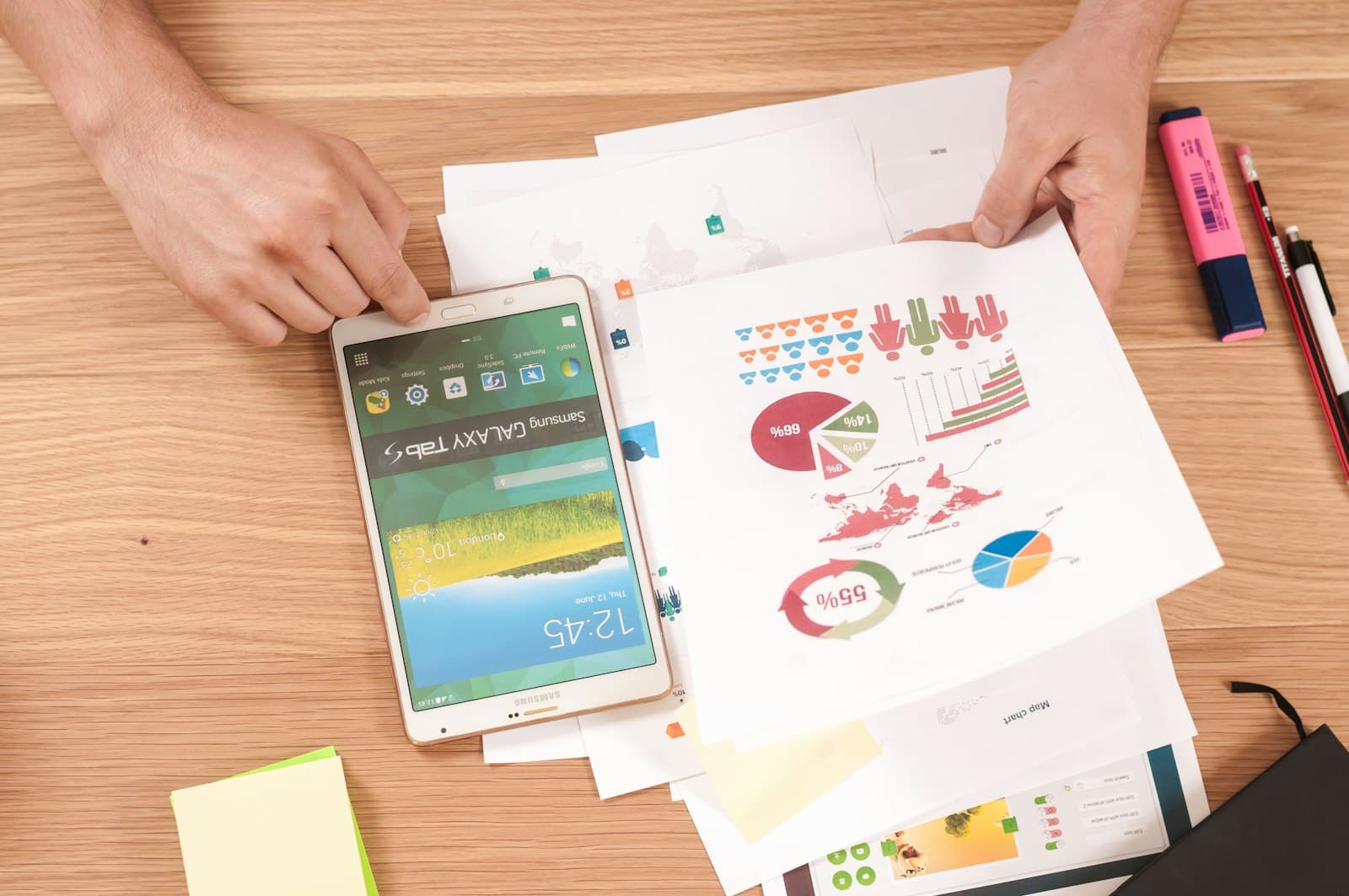 Hands holding printed charts and reports beside a tablet on a wooden desk.