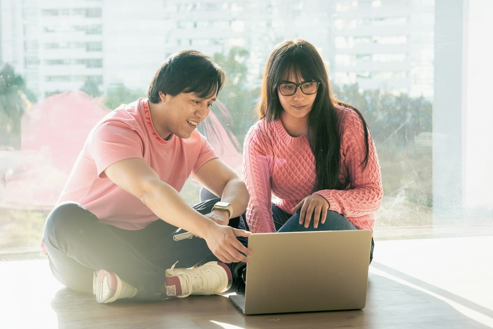 A couple sitting on the floor of a bright apartment, looking at a laptop together.